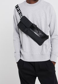 Borsa a tracolla nera con patta, pannello lucido e dettagli con zip, indossata sopra un maglione grigio. La tracolla regolabile presenta lettere bianche in grassetto.