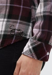 Plaid shirt with maroon, black, and white checks, cuff buttoned, "Soccx Spirit" embroidered in black near the hem, worn with black pants.