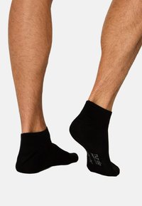Zwei Beine, die schwarze Knöchelsocken tragen, ein Fuß flach auf dem Boden und der andere leicht auf den Zehen erhoben.