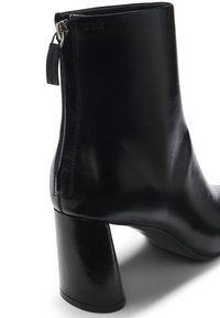 Bottine en cuir noir avec un talon géométrique épais, une fermeture éclair sur le côté et un logo en relief à l'arrière. Texture lisse, design épuré.