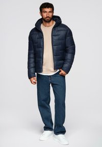 Giubbino puffo blu navy con cappuccio, design trapuntato, chiusura frontale con zip, indossato sopra una felpa beige, abbinato a jeans blu e sneakers bianche.
