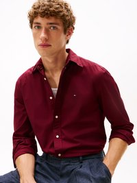 Bordeaux knoopshirt met een klein logo op de borst, lange mouwen opgerold, gecombineerd met grijze gemusterde broek. Gladde stoftextuur.