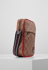 Sac brun à motifs avec garniture rouge, fermeture éclair et bandoulière réglable à rayures aux couleurs marine, rouge et crème. Patch du logo Fila sur le devant.