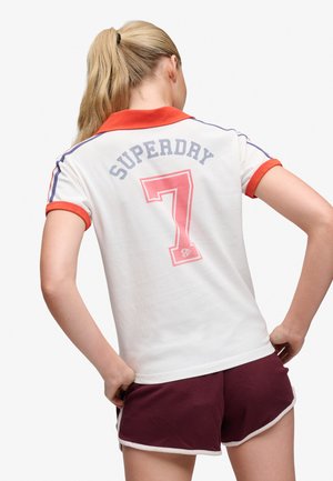 Frau mit blondem Pferdeschwanz, die ein weißes Polo mit rotem Kragen, mit dem Schriftzug "SUPERDRY 7" und bordeauxroten Shorts trägt, steht mit den Händen in der Hüfte und ist weggewandt.