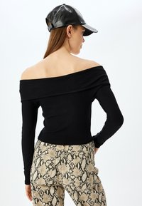Schwarzes, schulterfreies, geripptes Oberteil mit langen Ärmeln, kombiniert mit beigen Schlange-Muster-Hosen mit schwarzen Mustern. Eine schwarze Lederkappe wird getragen.