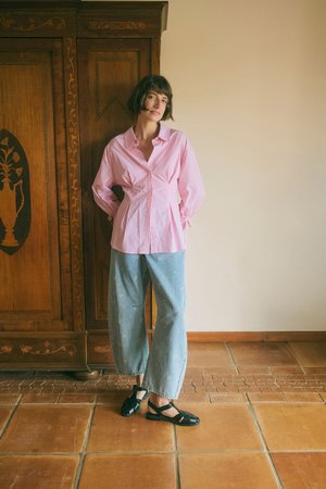 Femme aux cheveux courts debout à l'intérieur portant une chemise rose à rayures, un pantalon large bleu clair avec de petits motifs blancs et des sandales noires à bout fermé.