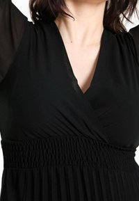 Robe noire avec un décolleté en V profond, des manches transparentes et une taille smockée avec des plis texturés ; tissu lisse avec une légère brillance.
