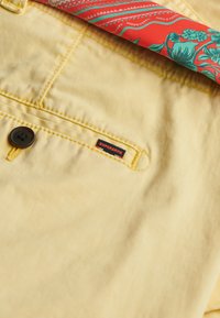 Tessuto giallo con una trama fine, un bottone nero e un'etichetta con la scritta "SUPERDRY". Un tessuto a fantasia rossa e verde si intravede.