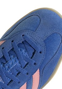 Μπλε σουέντ αθλητικά παπούτσια με ροζ ρίγες, διαθέτοντας υφαντές κορδόνια και λείο καπάκι από τη μύτη. Ο πάτος από καουτσούκ είναι ορατός στην κάτω πλευρά.