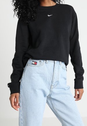 Kobieta nosząca czarną, długorękawową koszulkę Nike, wpuszczoną w jasnoniebieskie, wysokowalne spodnie dżinsowe Tommy Jeans.
