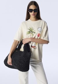 Zwarte geweven handtas met korte handvatten, met een gestructureerd oppervlak. Model draagt een lichtbeige graphic t-shirt en witte broek.