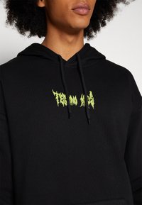 Sudadera negra de tejido suave con un bolsillo tipo canguro. Presenta un diseño gráfico en texto de color verde brillante en el pecho. Detalle de capucha con cordón.