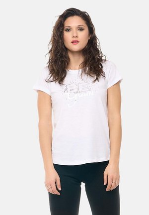 Femme aux cheveux bouclés portant un t-shirt blanc avec un motif graphique et un pantalon noir, debout devant un fond clair uni.