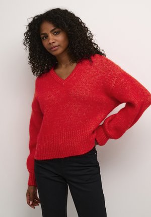 Maglione rosso a maglia con scollo a V, taglio morbido e polsini e orlo a coste. Caratterizzato da una texture morbida con una leggera lucentezza. Abbinato a pantaloni neri.