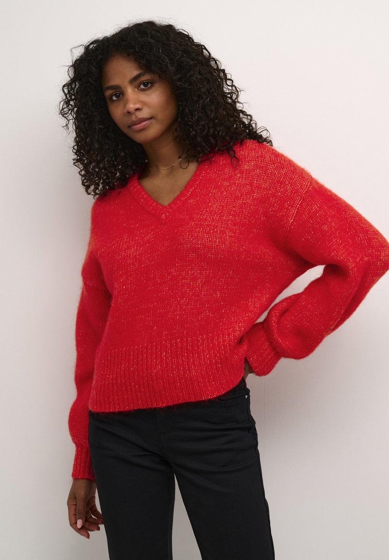 Maglione rosso a maglia con scollo a V, taglio morbido e polsini e orlo a coste. Caratterizzato da una texture morbida con una leggera lucentezza. Abbinato a pantaloni neri.