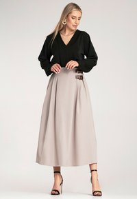 Czarna bluzka z długim rękawem i dekoltem w kształcie litery V; beżowa spódnica midi z podwójnymi brązowymi klamrami; czarne sandały na obcasie. Gładka tekstura materiału.
