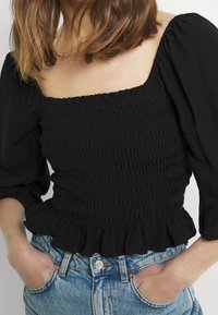 Černý nařasený top se širokými nafouklými rukávy a volánovým lemem, vyrobený z texturované látky; doplněný o modré džíny s vysokým pasem.