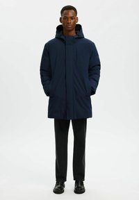 Cappotto con cappuccio blu navy caratterizzato da una zip frontale, tasche laterali e una texture leggermente imbottita, indossato sopra pantaloni scuri e scarpe.