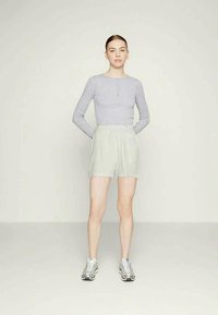Haut à manches longues gris clair avec des détails de boutons, associé à un short plissé vert clair. Le mannequin porte des chaussures de sport argentées, se tenant devant un fond uni.