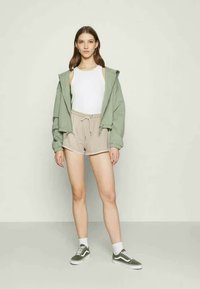 Hoodie verde oversized, top branco, calções bege, meias brancas e ténis verdes. O hoodie tem ombros caídos e um corte descontraído.