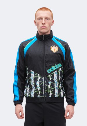 Jeune homme portant une veste de survêtement Adidas noire avec des touches bleues et un écusson 'League Cup Final 1992', debout devant un fond uni.