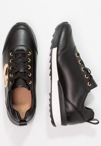Black athletic shoes made of smooth leather with gold eyelets, white midsole, and a textured black rubber sole. Features a logo on the side.