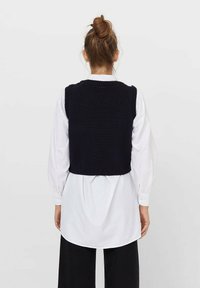 Kobieta z włosami w kok odkryta od tyłu, nosząca bezrękawnik w czarnym kolorze z dzianiny na długiej białej koszuli i czarnych spodniach, na jednolitym tle.