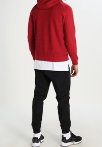 Červená mikina s kapucí a texturovanou tkaninou, položená na bílé košili, spárována s černými zúženými tepláky a černými teniskami s bílou podrážkou.