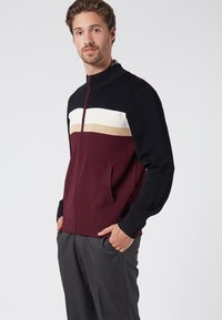 Strickjacke mit Reißverschluss, die einen schwarzen oberen Teil, einen weinroten Körper und horizontale Streifen in Beige und Weiß aufweist. Sie hat seitliche Taschen und gerippte Bündchen.