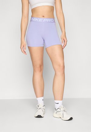 Mujer con pantalones cortos Nike Pro de color púrpura claro, sujetador deportivo blanco, calcetines Nike blancos y zapatillas de correr blancas, de pie sobre un fondo liso.