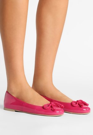 Des jambes portant des ballerines roses brillantes avec des nœuds en corde décoratifs, debout sur une surface blanche.