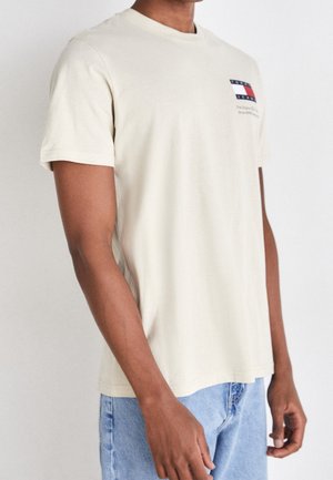 T-shirt en coton couleur crème avec un col rond, des manches courtes, et un petit patch drapeau sur la poitrine gauche. Coupe standard, texture douce.