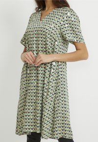 Grön och beige klänning med V-hals, korta ärmar och ett geometriskt triangelmönster i marinblått och grönt. Lättviktigt tyg med en avslappnad passform.