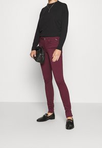 Jeans skinny burdeos con una cintura de altura media, combinados con una blusa negra de manga larga y mocasines negros. Sosteniendo un pequeño bolso negro.