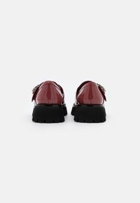 Glänzende bordeauxfarbene Lacklederschuhe mit einer klobigen schwarzen Sohle, ausgestattet mit dekorativen Hardware-Details an der Rückseite und einer abgerundeten Silhouette.