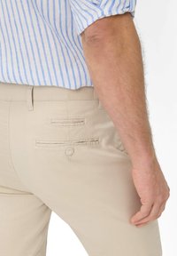 Pantaloni beige con una finitura texturizzata. Presentano due tasche posteriori con chiusura a bottone e passanti per cintura. Accompagnati da una camicia a righe blu chiaro.