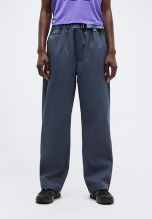 Persoană purtând pantaloni largi albaștri închiși cu o curea albastru deschis, pantofi negri și un top mov, stând în fața unui fundal simplu și deschis la culoare.