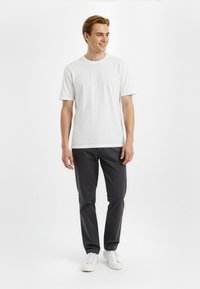 White short-sleeve t-shirt, crew neck, paired with dark gray pants and white sneakers, standing against a plain background.