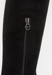 Zwarte suède laars schacht met een verticale zwarte rits en een ronde zilveren ritstrekker op een witte achtergrond.