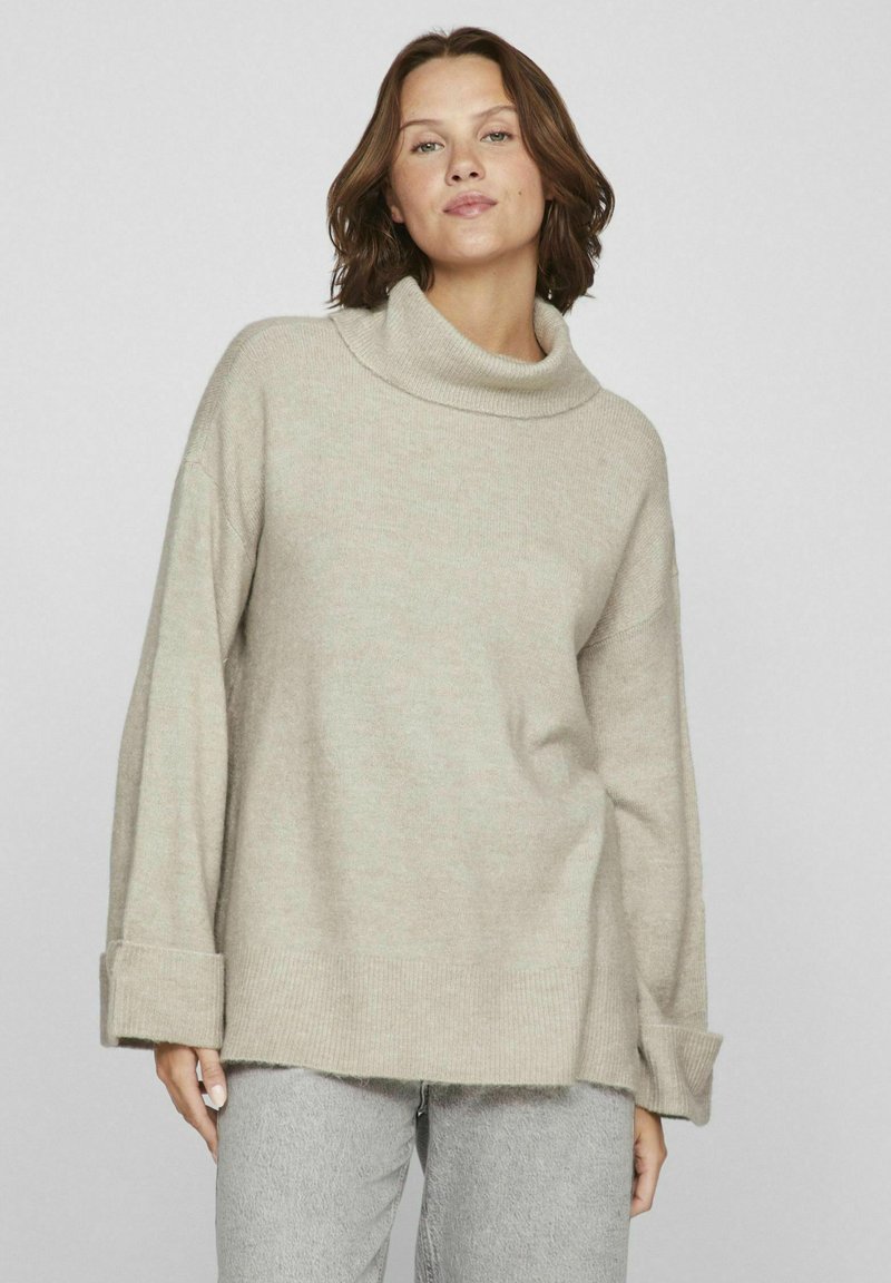 Pull en tricot beige avec un large col bénitier, des épaules tombantes et des poignets reversés. Texture douce avec une coupe décontractée, associé à un jean gris.