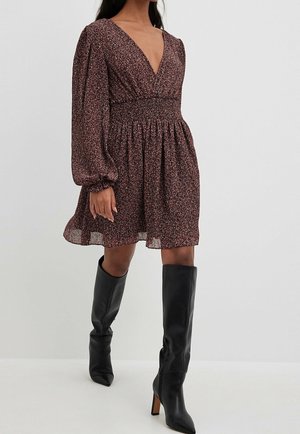 Femme portant une robe fleurie à encolure en V profonde avec de longues manches bouffantes, associée à des bottes noires montantes à talons, debout devant un fond uni.