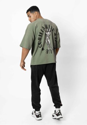 Man draagt een oversized olijfgroen overhemd met een zwarte gespierde gorilla grafiek en tekst, zwarte broek en witte sneakers, staand naastwaarts.