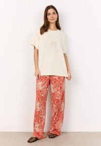 Cream oversized t-shirt with palm print graphic. Loose-fit red pants in a white wave pattern. Textured slide sandals complete the outfit.