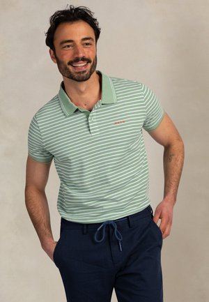 Glimlachende man met donker haar en baard, gekleed in een groen-wit gestreept poloshirt en marineblauwe broek met trekkoord, staand met handen in de zakken.