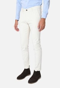Pantaloni slim-fit di colore chiaro in cotone, con vita piatta, tasche laterali e chiusura con bottone marrone. Indossati con stivali in pelle scamosciata scuri.