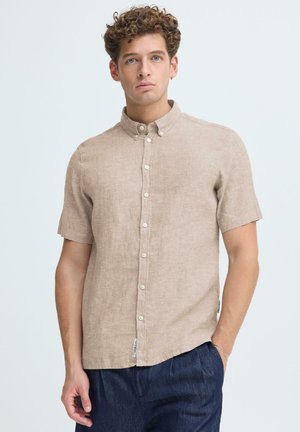 Giovane con capelli ricci che indossa una camicia beige a maniche corte con bottoni e pantaloni blu scuro, in piedi con una mano in tasca.