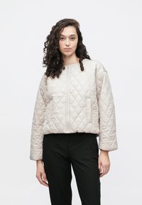 Beżowa pikowana kurtka o pudełkowym kroju, z okrągłym dekoltem i zamkiem błyskawicznym z przodu. Charakteryzuje się ukośnym przeszyciem i bocznymi kieszeniami, zestawiona z czarnymi spodniami.