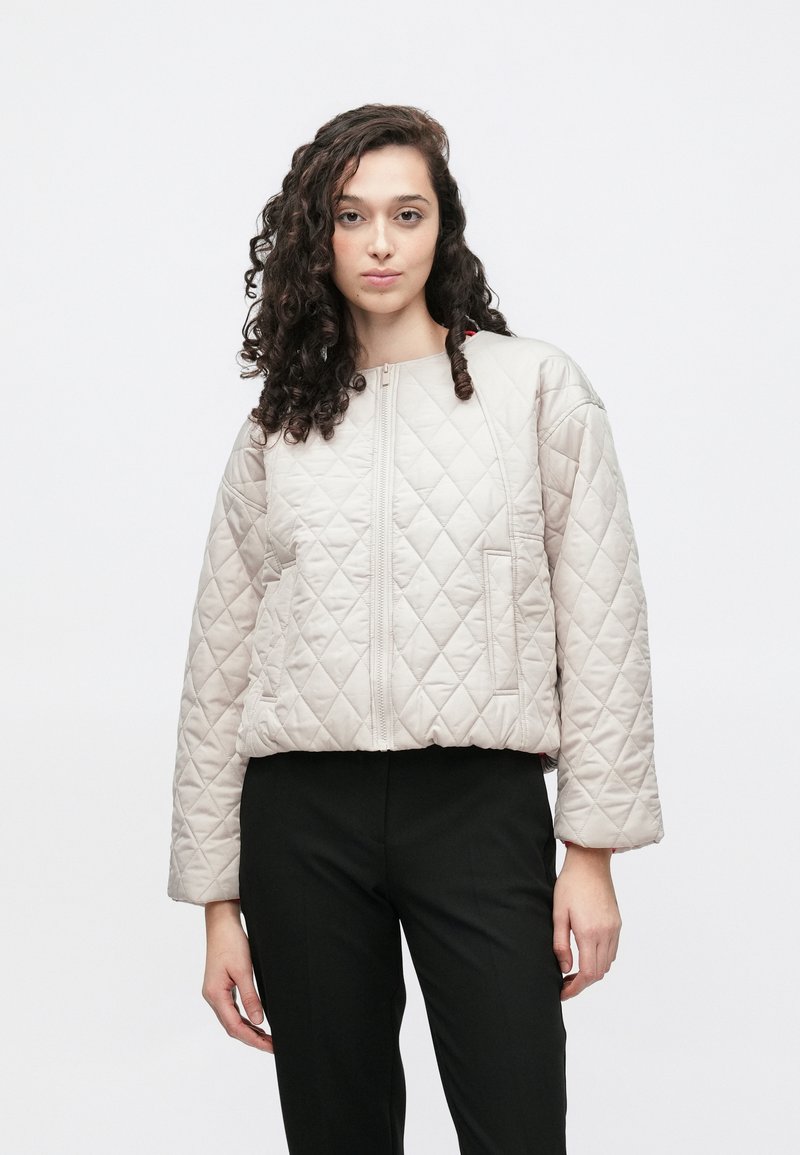 Beżowa pikowana kurtka o pudełkowym kroju, z okrągłym dekoltem i zamkiem błyskawicznym z przodu. Charakteryzuje się ukośnym przeszyciem i bocznymi kieszeniami, zestawiona z czarnymi spodniami.