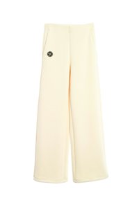 Pantalones de pierna ancha en tejido crema, con una textura suave, dos bolsillos laterales y un parche negro con el logo en la parte frontal.