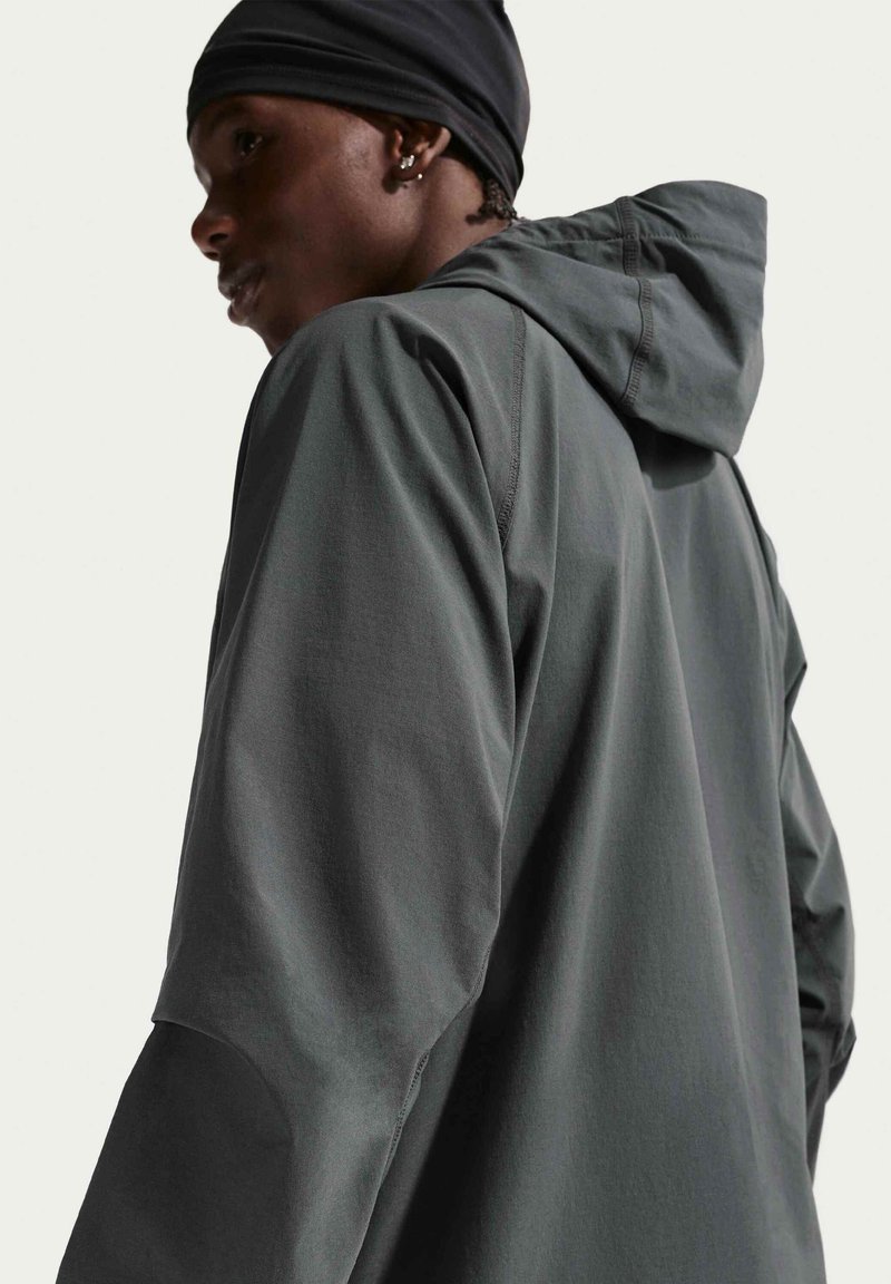 Person wearing a dark gray hooded jacket and black headwrap, facing left with a neutral background.